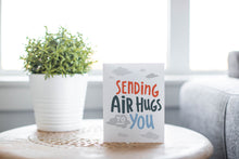 Load image into Gallery viewer, A greeting card laying on a wooden table with some cut wood details. The card features the words “Sending air hugs to you.”