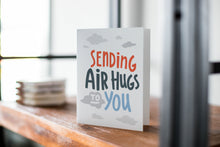Load image into Gallery viewer, A card on a wood tabletop with an object in the background that is out of focus. The card features the words “Sending air hugs to you.”
