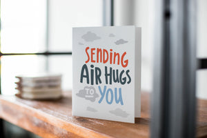 A card on a wood tabletop with an object in the background that is out of focus. The card features the words “Sending air hugs to you.”