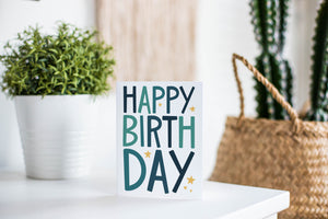 A greeting card is featured on a white tabletop with a white planter in the background with a green plant. There’s a woven basket in the background with a cactus inside. The card features the words “Happy birthday” with blue letters featured on a white background.