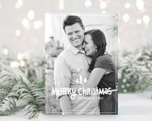 Load image into Gallery viewer, A photo of a one-sided Christmas card showing the front of the card standing up with pine needles behind and blurred white Christmas lights. The photo card features one photo with the words “Merry Christmas” with illustrated pine trees above the words. There’s a family name under the words which you can edit with your name.