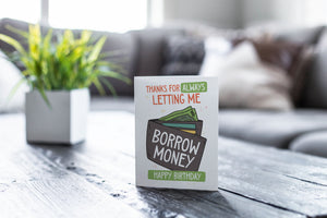 A greeting card featured on a black, wood coffee table. There’s a white planter in the background with a green plant. There’s also a gray sofa in the background with a white pillow. The card features the words “Thanks for always letting me borrow money, happy birthday!” with an illustrated wallet.
