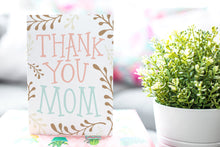 Load image into Gallery viewer, A photo of a card featured on a tabletop next to a white planter filled with a green plant. ​​The card features the words “Thank You Mom” with illustrated plant leaves.