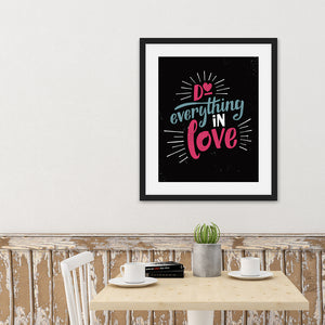 A black art print in a black frame hangs on a cafe wall. Below it is a small table holding coffee cups, books, and a plant. The print reads "Do everything in love" in bright pink and blue hand-lettering style, with white dashes around the words.