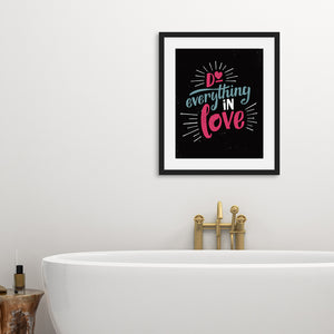 A black art print in a black frame hangs on a white bathroom wall. Below it is a freestanding white bath with brass taps.  The print reads "Do everything in love" in bright pink and blue hand-lettering style, with white dashes around the words.