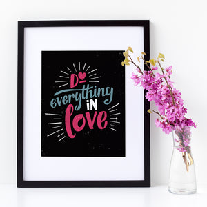 A small black art print in a black frame leans against a white wall. Beside it is a vase of pink flowers. The print reads "Do everything in love" in bright pink and blue hand-lettering style, with white dashes around the words.