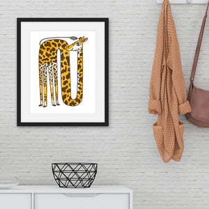 A black frame on a wall with a yellow giraffe illustration. The frame is on the wall above a white side table and next to a coat rack. 