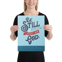 Load image into Gallery viewer, A woman is holding an art canvas in front of her and smiling. The print is bright blue with the verse &#39;Be Still and Know that I am God&#39; illustrated in a bold typographic style. The woman is white with blond hair and wearing black.