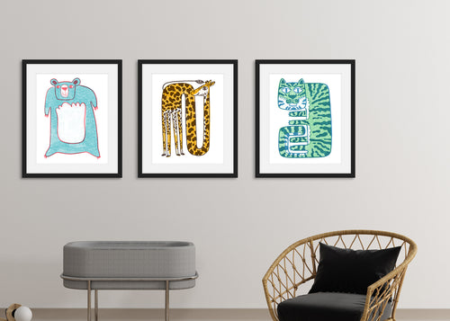 Three black frames on a white wall above a baby cot and chair. The black frames have a blue bear illustration, a giraffe illustration and a a tiger illustration. 