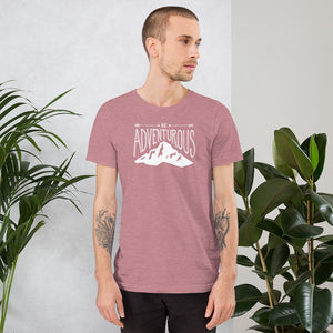 A man wearing an orchid pink short sleeved t-shirt. The tee features the lettering and illustration in white. The phrase “Be adventurous” with arrows pointing to the word “be” and a mountain illustration underneath the word “adventure.”