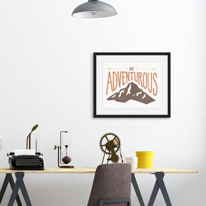 A black frame featured on the wall above a desk. The artwork in the frame says “Be adventurous” with arrows pointing to the word “be” and a brown mountain illustration underneath the word “adventure.”