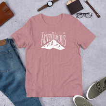 Load image into Gallery viewer, An orchid pink T-shirt laying flat with objects around it. The tee features the lettering and illustration in white. The phrase “Be adventurous” with arrows pointing to the word “be” and a mountain illustration underneath the word “adventure.”