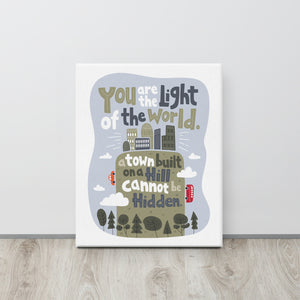 A canvas is leaning against a wall on the floor. The artwork is on a white background with lettering reading "You are the light of the world, a town built on a hill cannot be hidden." The words are a light gray background with an illustrated city.