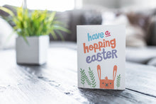 Load image into Gallery viewer, A greeting card featured on a black, wood coffee table. There’s a white planter in the background with a green plant. There’s also a gray sofa in the background with a white pillow. The card features an illustrated Easter bunny with some leaves around the bunny. The words “have a hopping Easter” are above the bunny. 