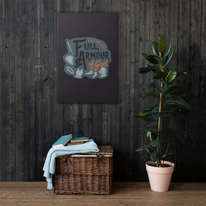 A dark gray canvas hangs on a dark wooden-clad wall, over a wicker basket holding books and blankets and a large potted plant. The canvas features the quote 'Put on the full armour of God' in black and orange typography, with medieval-style pieces of armour illustrated in pale gray.