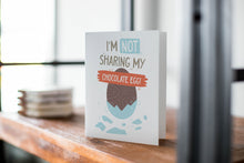 Load image into Gallery viewer, A card on a wood tabletop with an object in the background that is out of focus. The card features an illustrated chocolate Easter egg with the words “I’m not sharing my chocolate egg!”