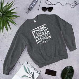 A dark grey sweatshirt laying on the ground with objects around it. The sweatshirt features hand drawn lettering in white with the words "Our creativity is an outflow of His."