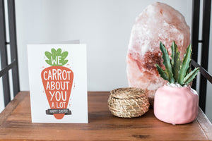 A card on a wood tabletop and on the right side of the card is a woven basket, a pink plant pot with a cactus in it and a pink crystal rock. The card features the words “We carrot about you a lot, Happy Easter,” all featured in an illustrated carrot.