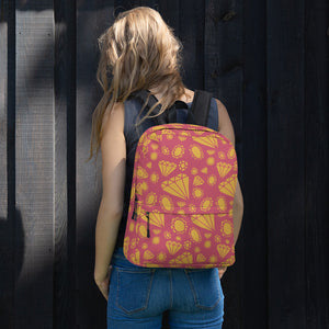 A woman standing in front of a black fence with her back to the camera. She has a backpack over both shoulders, resting on her back.  The backpack features illustrated gems and diamonds in gold on top off a muted hot pink colored backpack. 