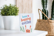 Load image into Gallery viewer, A greeting card is featured on a white tabletop with a white planter in the background with a green plant. There’s a woven basket in the background with a cactus inside. The card features an illustrated Easter bunny with some leaves around the bunny. The words “have a hopping Easter” are above the bunny. 