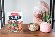 Load image into Gallery viewer, A card on a wood tabletop and on the right side of the card is a woven basket, a pink plant pot with a cactus in it and a pink crystal rock. The card features the words “Happy Easter” with illustrated Easter eggs.