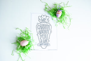 A coloring sheet on a white tabletop. There’s fake Easter grass around the color page. The card features the words “We carrot about you a lot, Happy Easter,” all featured in an illustrated carrot.