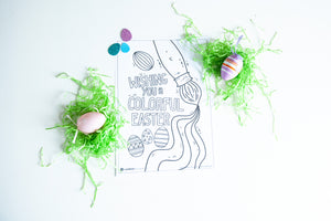 A coloring sheet on a white tabletop. There’s fake Easter grass around the color page. The page features an illustrated paint brush and Easter eggs with the words “Wishing you a colorful Easter.”