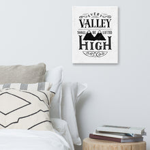 Load image into Gallery viewer, A monochrome art canvas hangs on a pale grey bedroom wall. The canvas has a white background, with the words &#39;Every valley shall be lifted high&#39; in black typographic lettering with flourishes and an image of two mountain peaks. Beneath the canvas there’s a bed with lots of white pillows and a wooden side table supporting a pile of books.