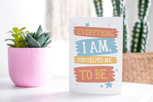A greeting card featured standing up on a white tabletop with a pink plant pot in the background and some succulents in the pot. There’s a woven basket in the background with a cactus inside. The card features the words “Everything I Am, You Helped Me To Be.”