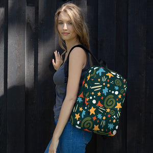 A woman standing by a black fence with one hand on the fence. She is turned sideways with a backpack over one arm. The backpack is hunter green with a fun pattern of yellow stars, green swirls, blue "splats" and other fun whimsical shapes. The backpack straps are black. 