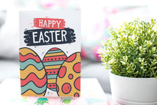 Load image into Gallery viewer, A photo of a card featured on a tabletop next to a white planter filled with a green plant. ​​The card features the words “Happy Easter” with illustrated Easter eggs in muted bright colors.