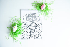 A coloring sheet on a white tabletop with fake green Easter grass and Easter eggs around the sheet. The coloring sheet features the words “Happy Easter” with illustrated Easter eggs. 