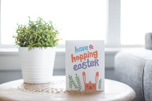 A greeting card laying on a wooden table with some cut wood details. The card features an illustrated Easter bunny with some leaves around the bunny. The words “have a hopping Easter” are above the bunny. 