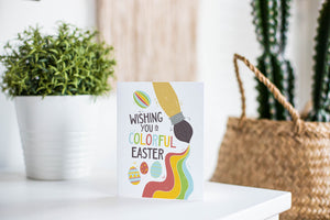 A greeting card is featured on a white tabletop with a white planter in the background with a green plant. There’s a woven basket in the background with a cactus inside. The card features an illustrated paint brush and Easter eggs with the words “Wishing you a colorful Easter.”