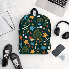 Load image into Gallery viewer, A backpack is placed on a table with a laptop, notebook, shoes, headphones and a mobile phone. The backpack is hunter green with a fun pattern of yellow stars, green swirls, blue &quot;splats&quot; and other fun whimsical shapes. The backpack straps are black. 