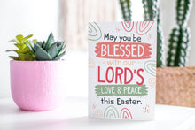 Load image into Gallery viewer, A greeting card featured standing up on a white tabletop with a pink plant pot in the background and some succulents in the pot. There’s a woven basket in the background with a cactus inside. The card features the words “May You be Blessed with our Lord&#39;s Love &amp; Peace this Easter.”