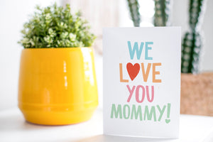 A greeting card is on a table top with a yellow plant pot and a green plant inside. The card features the words “We Love You Mommy!” 