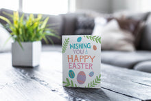 Load image into Gallery viewer, A greeting card featured on a black, wood coffee table. There’s a white planter in the background with a green plant. There’s also a gray sofa in the background with a white pillow. The card features the words “Wishing you a happy Easter” with illustrated Easter eggs and palm leaves.