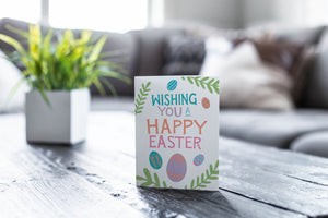 A greeting card featured on a black, wood coffee table. There’s a white planter in the background with a green plant. There’s also a gray sofa in the background with a white pillow. The card features the words “Wishing you a happy Easter” with illustrated Easter eggs and palm leaves.