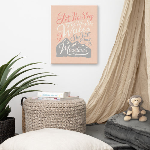 An illustrated pink canvas hangs on a white wall, next to a fabric canopy and children's books and toys. The canvas reads 'Let her sleep for when she wakes she will move mountains' in a pink, white, and light grey lettering design, with a grey mountain illustration at the bottom.
