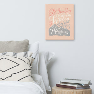 An illustrated pink canvas hangs on a white wall, over a white bed and a stack of books on a bedside table. The canvas reads 'Let her sleep for when she wakes she will move mountains' in a pink, white, and light grey lettering design, with a grey mountain illustration at the bottom.
