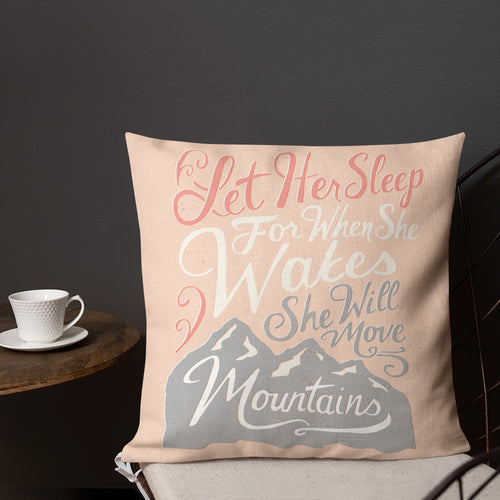 A pink cushion sits on a chair against a dark wall. The cushion reads 'Let her sleep for when she wakes she will move mountains' in a pink, white and light grey lettering design, with a grey mountain illustration
