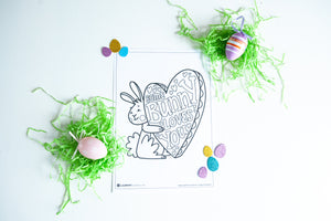 A coloring sheet on a white tabletop. There’s fake Easter grass around the color page. The card features an illustrated Easter bunny holding a heart with the words “some bunny loves you.” The illustrations and words are able to be colored in. 