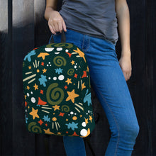 Load image into Gallery viewer, A woman leaning on a black fence with just her hands, waist and jeans showing. She is holding a backpack by its top loop strap. The backpack is hunter green with a fun pattern of yellow stars, green swirls, blue &quot;splats&quot; and other fun whimsical shapes. The backpack straps are black. 