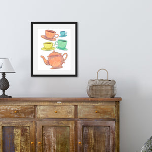 A black frame is hanging above a brown credenza with a lamp and basket on top of it. The frame contains artwork on a white background with four teacups on saucers and one large teapot. The teacups are in muted colors of orange, blue, yellow and green and the teapot is a muted orange. On the teacups, saucers and teapot there is a light flower detail pattern.
