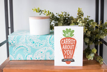 Load image into Gallery viewer, A greeting card is on a table top with a present in blue wrapping paper in the background. On top of the present is a candle and some greenery from a plant too. The card features the words “We carrot about you a lot, Happy Easter,” all featured in an illustrated carrot. 