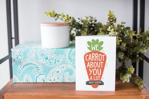 A greeting card is on a table top with a present in blue wrapping paper in the background. On top of the present is a candle and some greenery from a plant too. The card features the words “We carrot about you a lot, Happy Easter,” all featured in an illustrated carrot. 