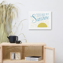Load image into Gallery viewer, A white canvas hangs on a white wall over a wooden cane cabinet. The print reads &#39;You are my sunshine&#39; in blue over a yellow sun illustration