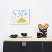 Load image into Gallery viewer, A white canvas hangs on a white wall over a black kitchen cupboard and wooden utensils. The canvas reads &#39;You are my sunshine&#39; in blue over a yellow sun illustration