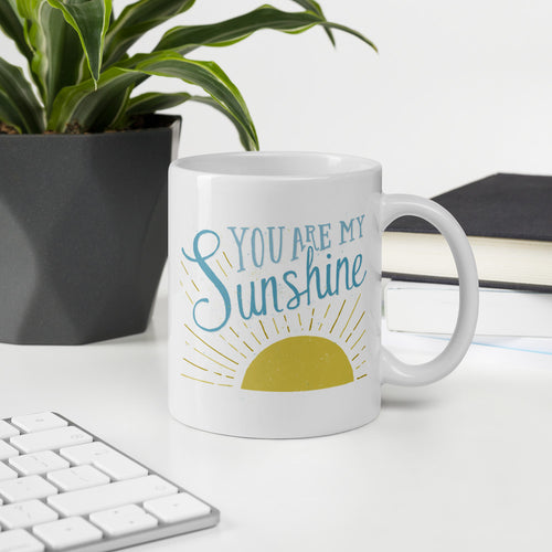 A white mug sits on a white desk beside a potted plant.  The mug reads 'You are my sunshine' in blue over a yellow sun illustration